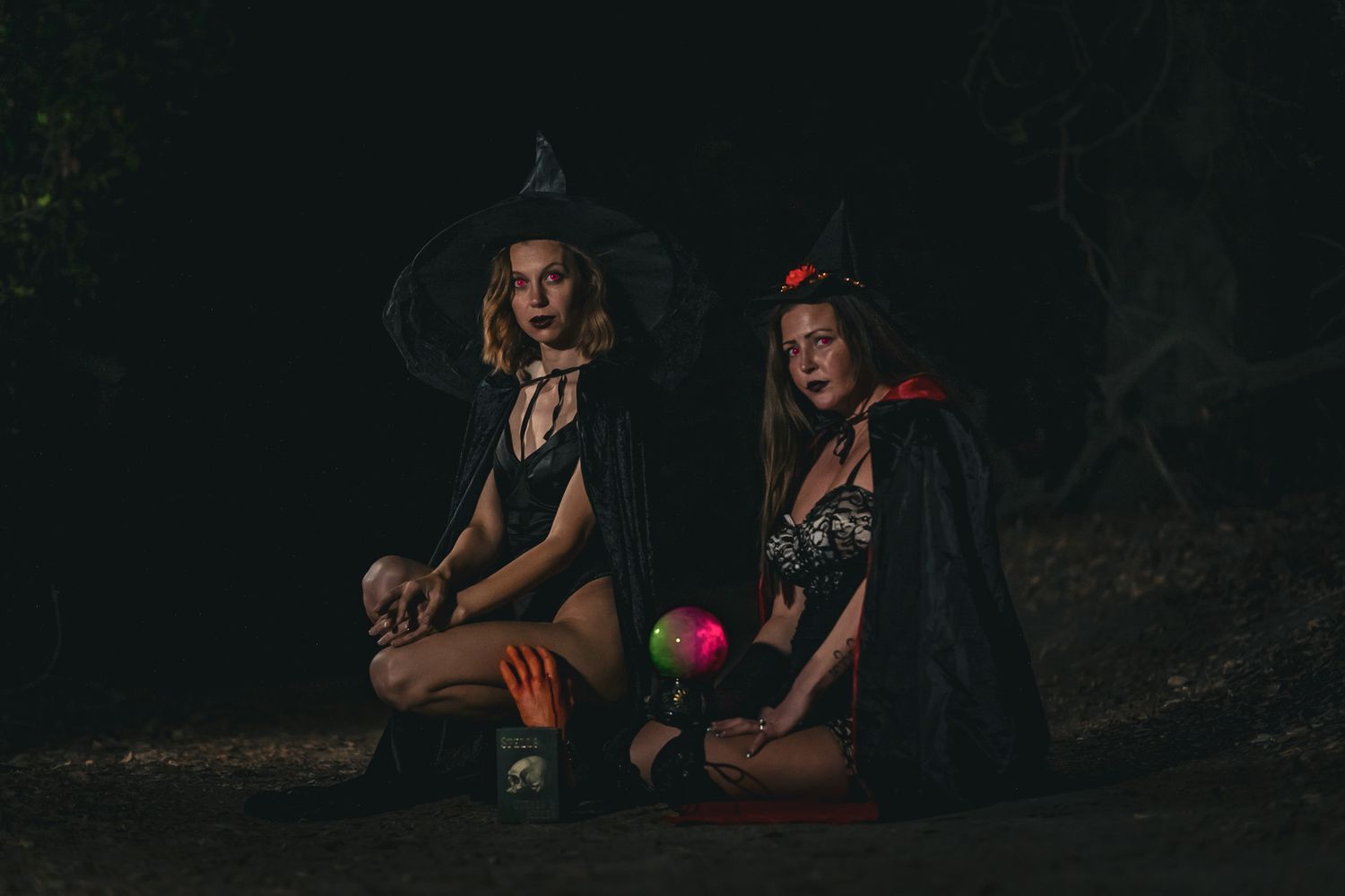Two figures in witch costumes sitting in a dark forest with glowing orbs and pumpkins.