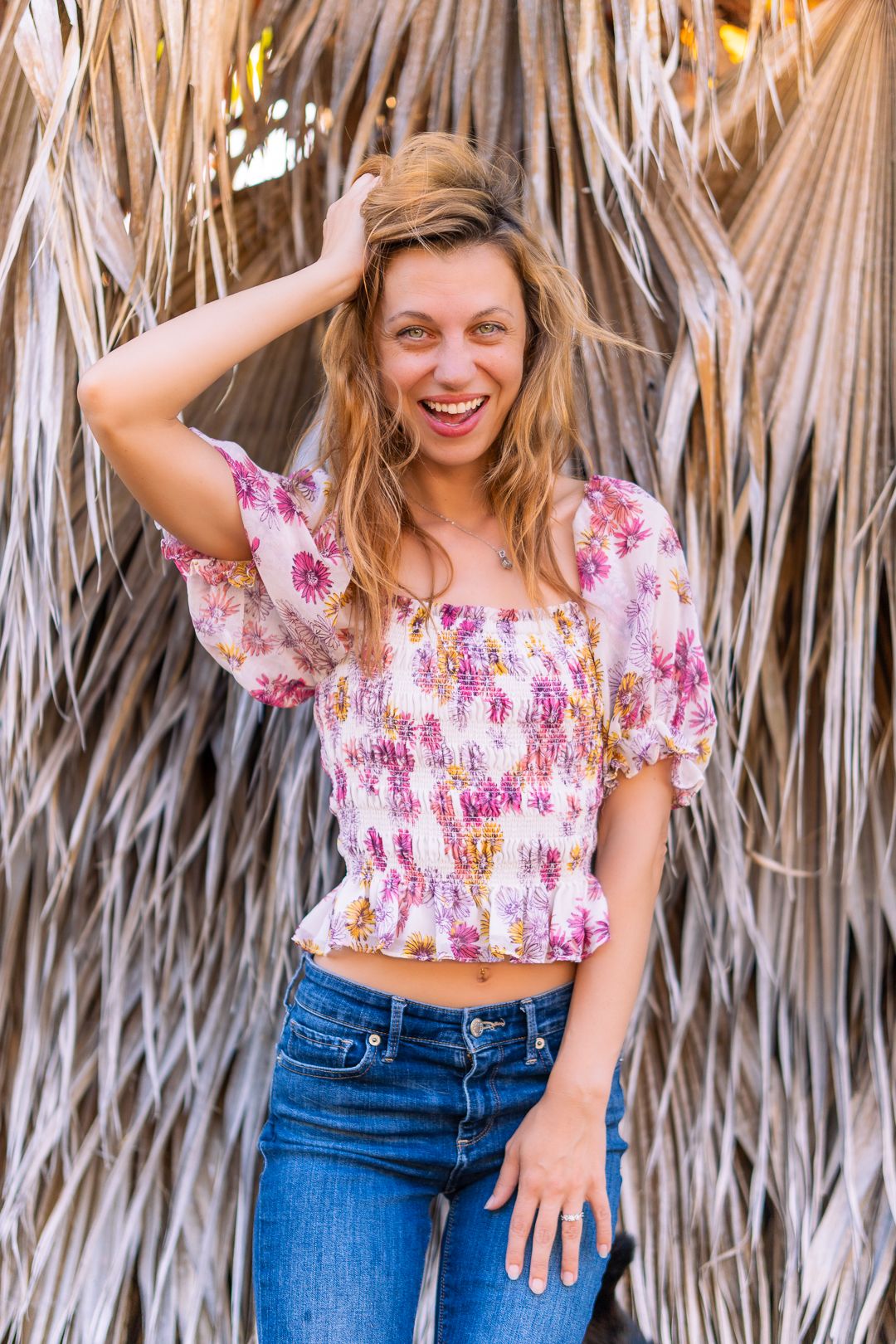 Candid lifestyle photo of someone laughing in floral top and jeans against palm frond background.