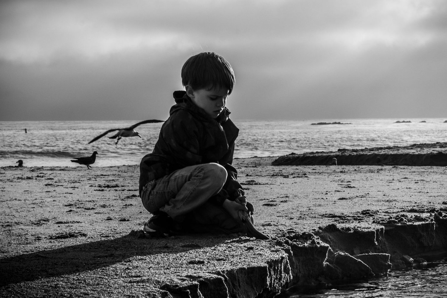 A silhouette sits in contemplation on a beach during an overcast day in black and white.