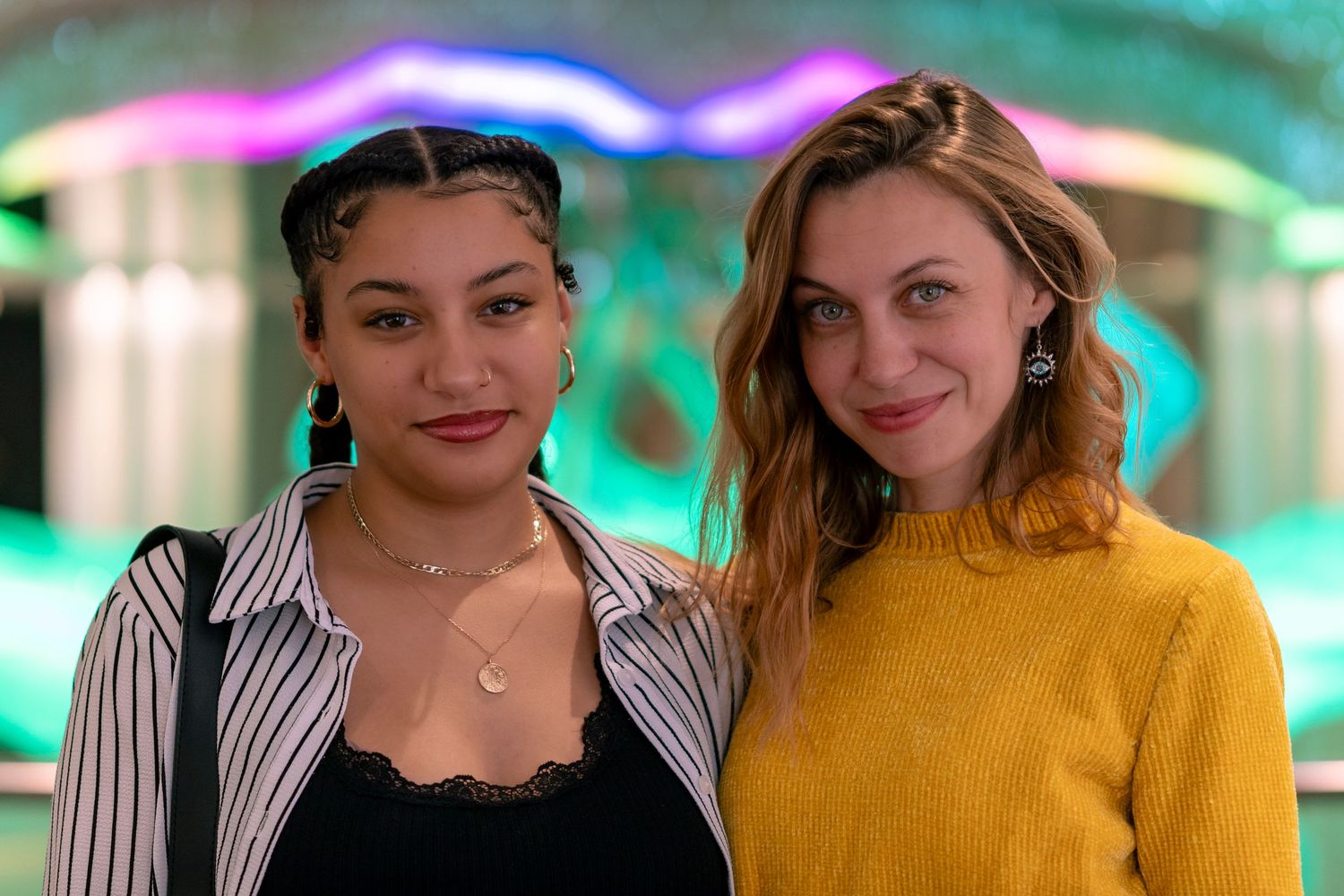 Two people stand together indoors, one wearing a striped shirt and one in a yellow sweater, with colorful lighting behind them.