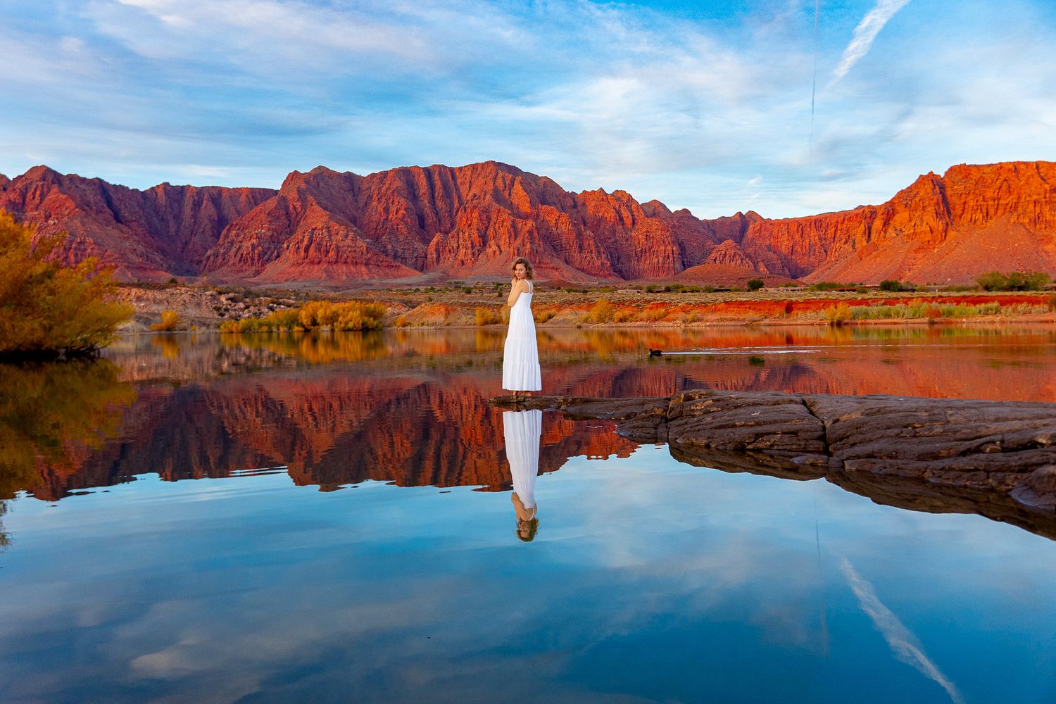 Red rock cliffs reflect perfectly in calm lake waters with a small sailboat in Utah's desert landscape.