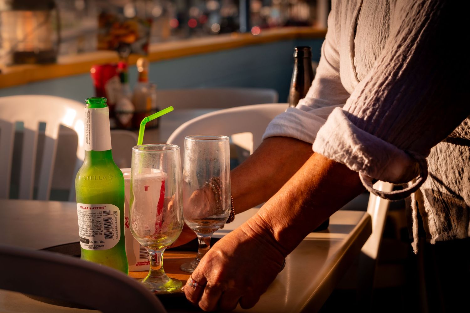 Hand pouring drinks from bottles into glasses at a dimly lit bar counter with empty glassware and bottles.