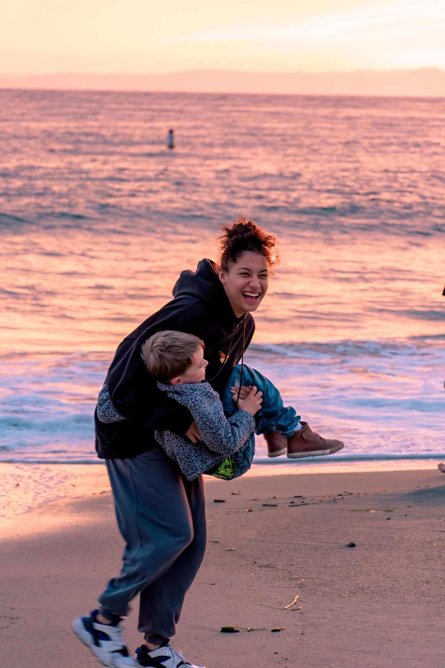 Two people share a joyful moment playing and laughing together on a beach at sunset with pink and purple hues in the sky.
