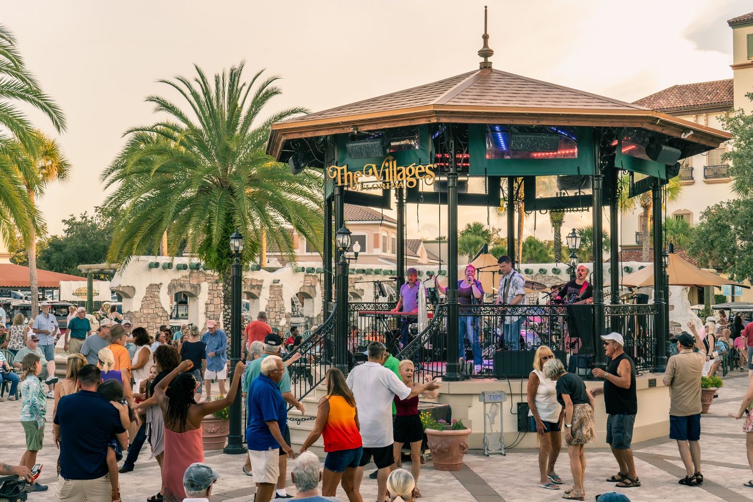 A crowd gathers around a gazebo bandstand with palm trees at sunset during an outdoor concert performance.