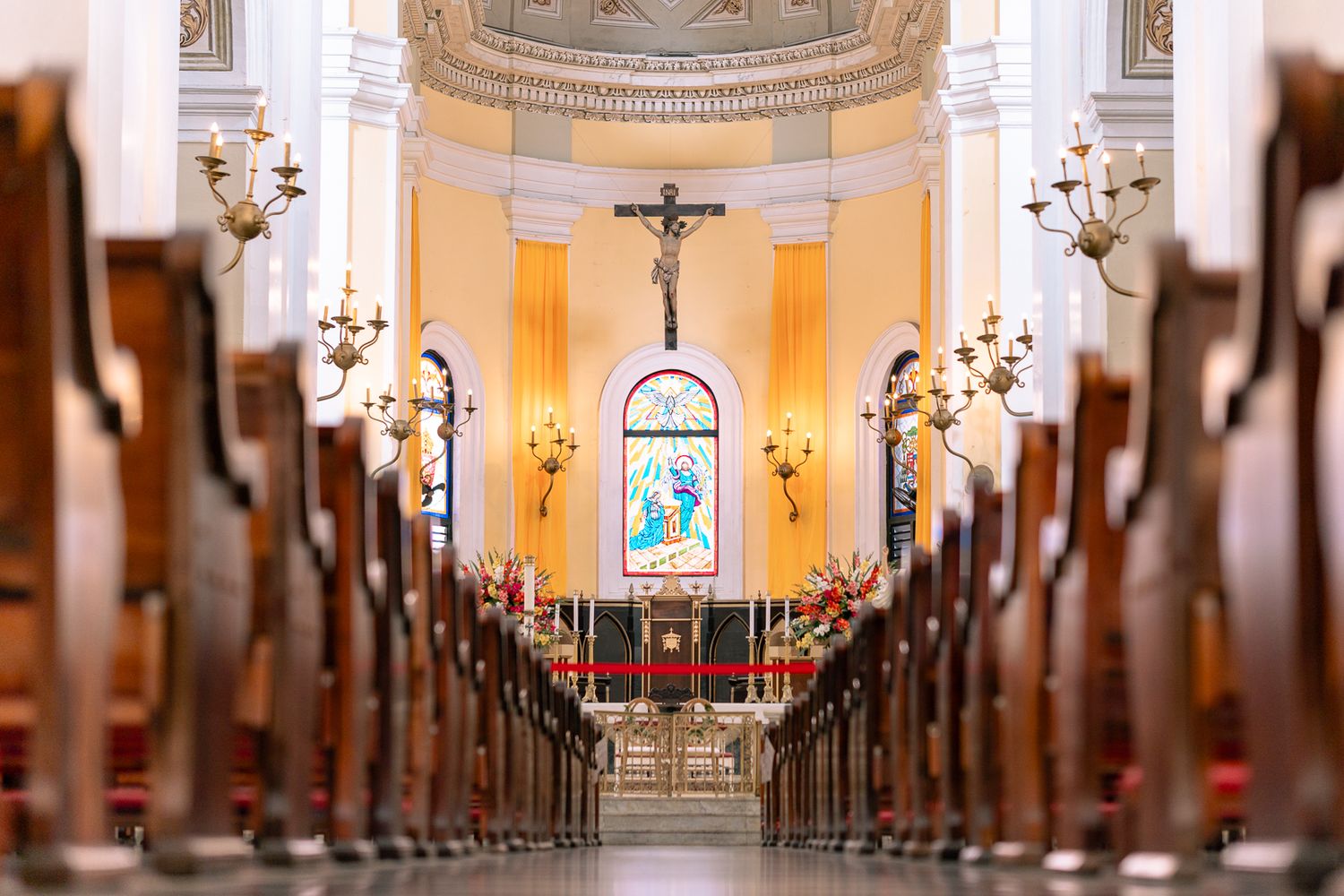 Church interior with wooden pews leading to an altar with a crucifix and stained glass window in soft focus.