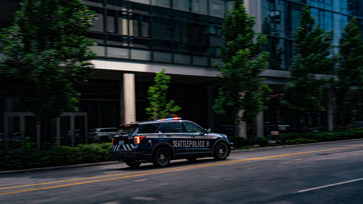 Black police patrol vehicle driving on urban street with trees and modern buildings in background at dusk.