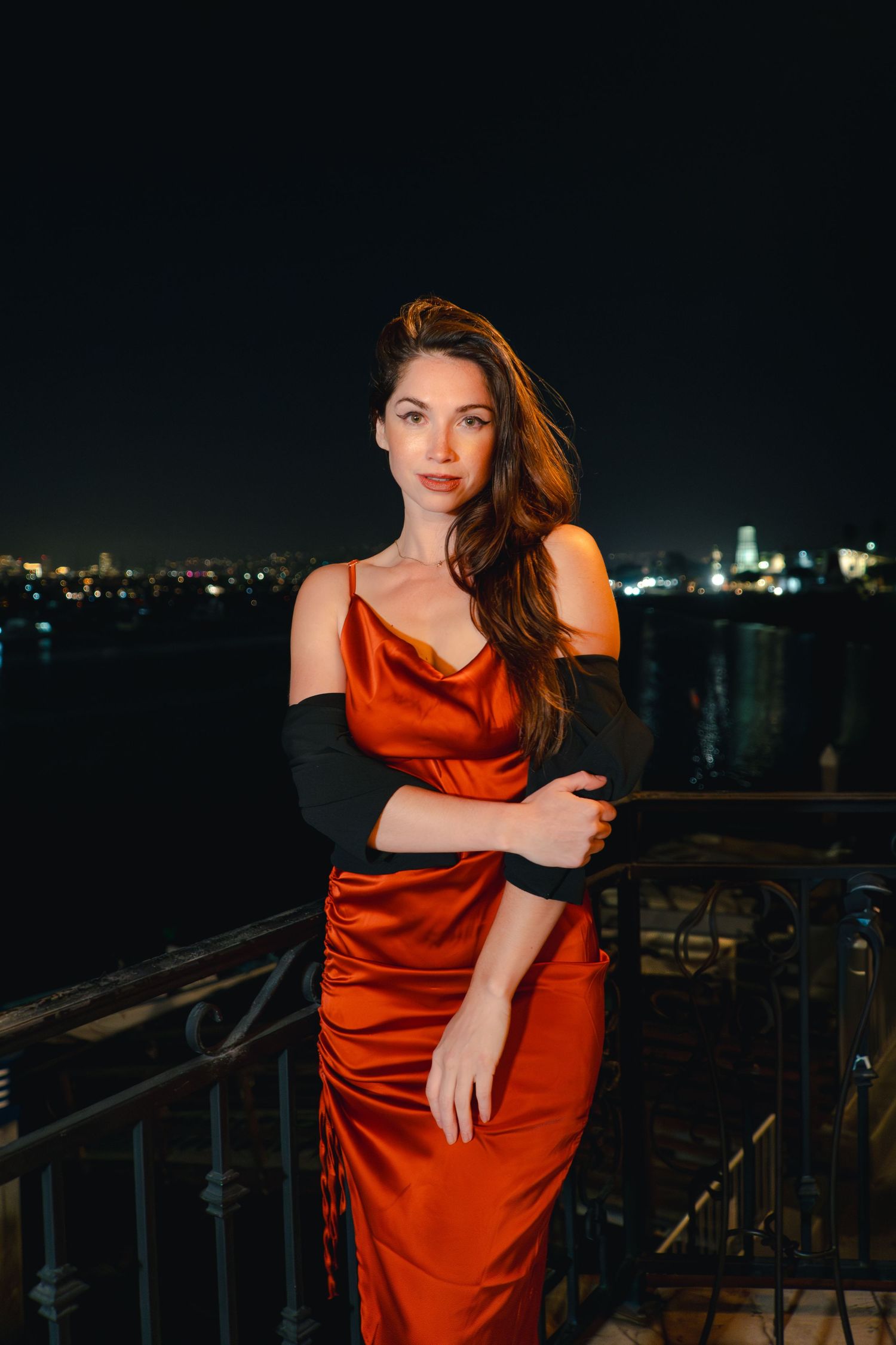 Glamorous nighttime rooftop portrait in red silk dress against city lights.