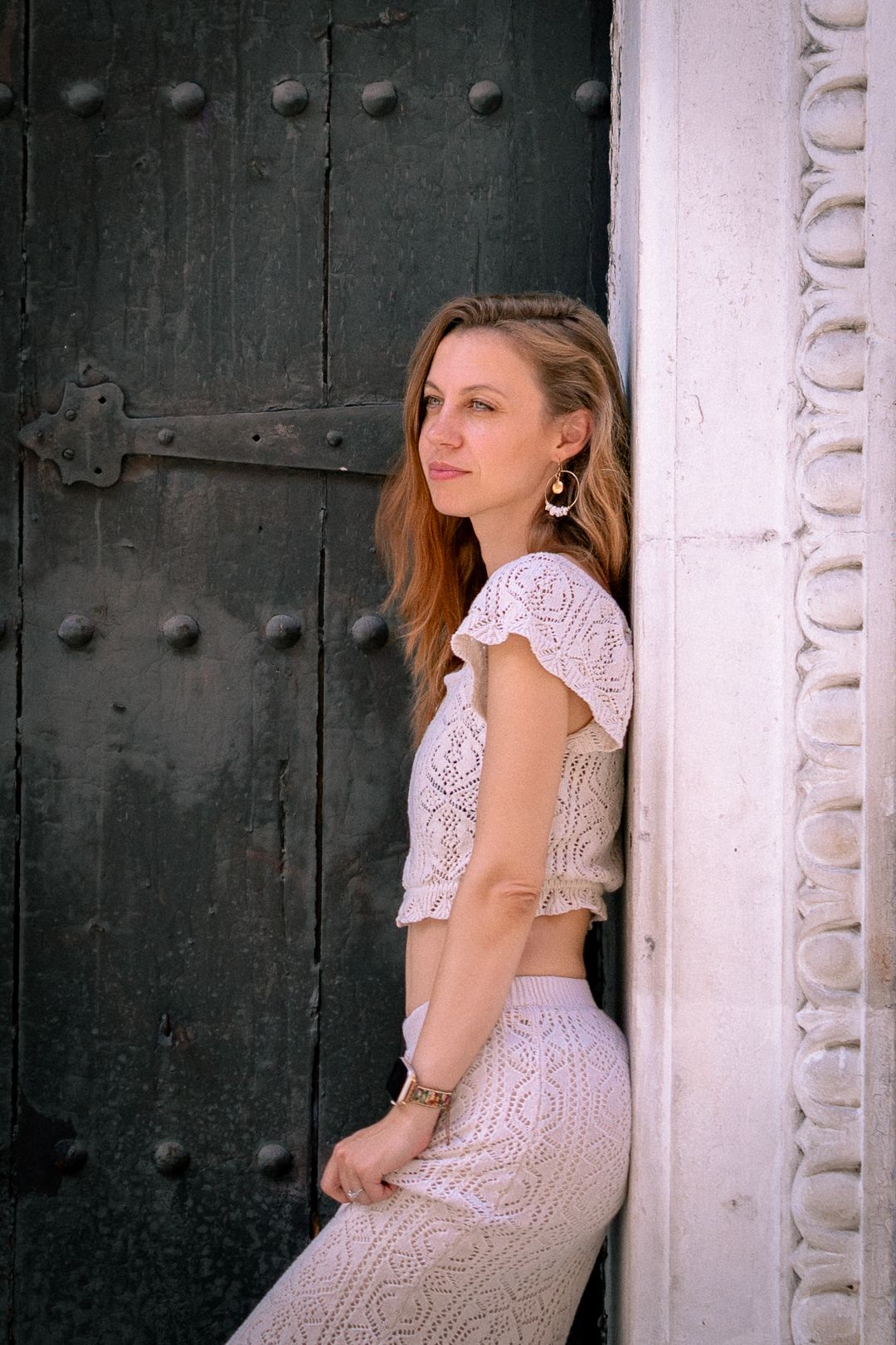 Fashion portrait in white lace outfit posed against dark wooden door in architectural setting.