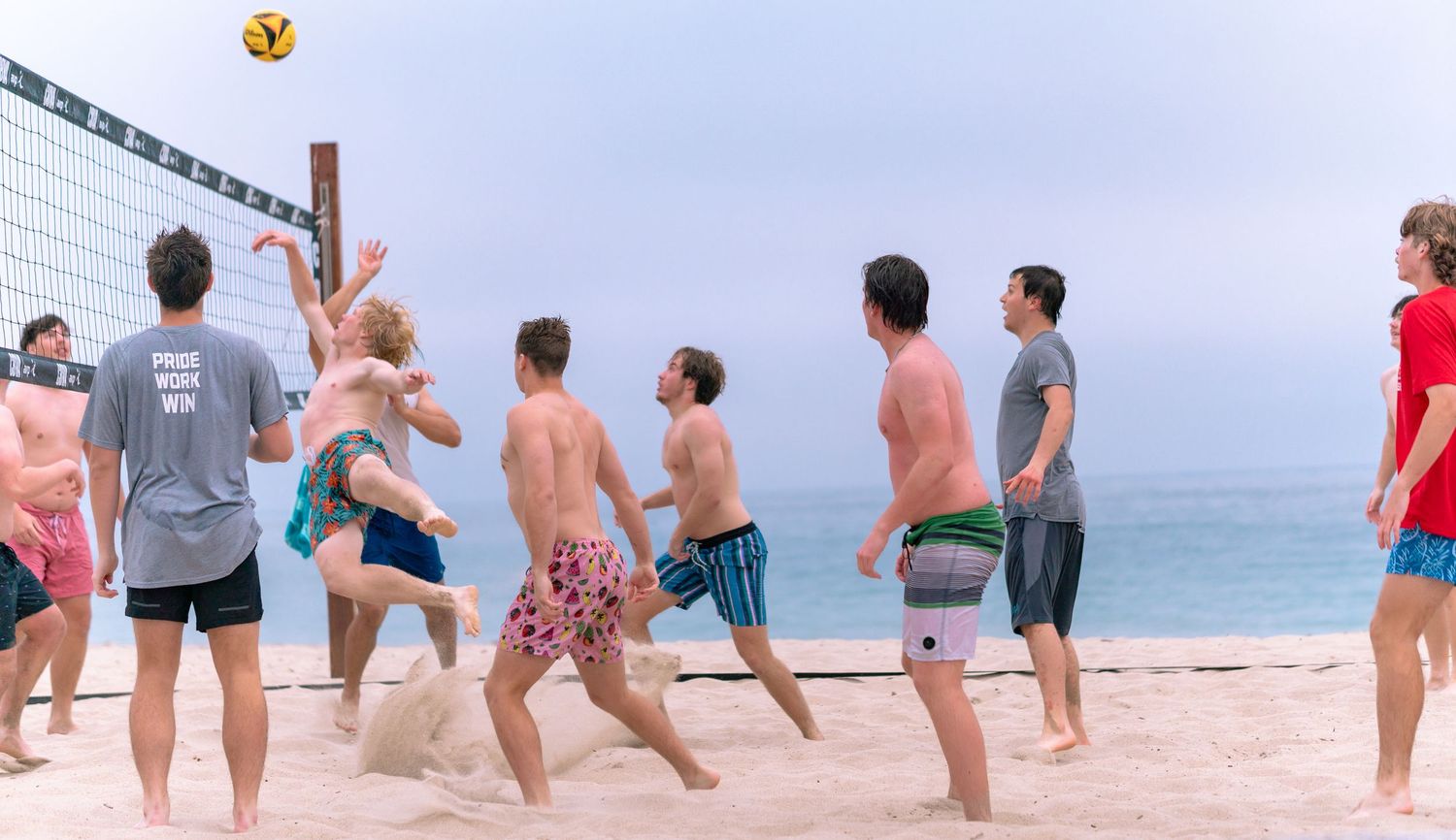 A group of people playing beach volleyball on a sandy shore with the ocean visible in the background.