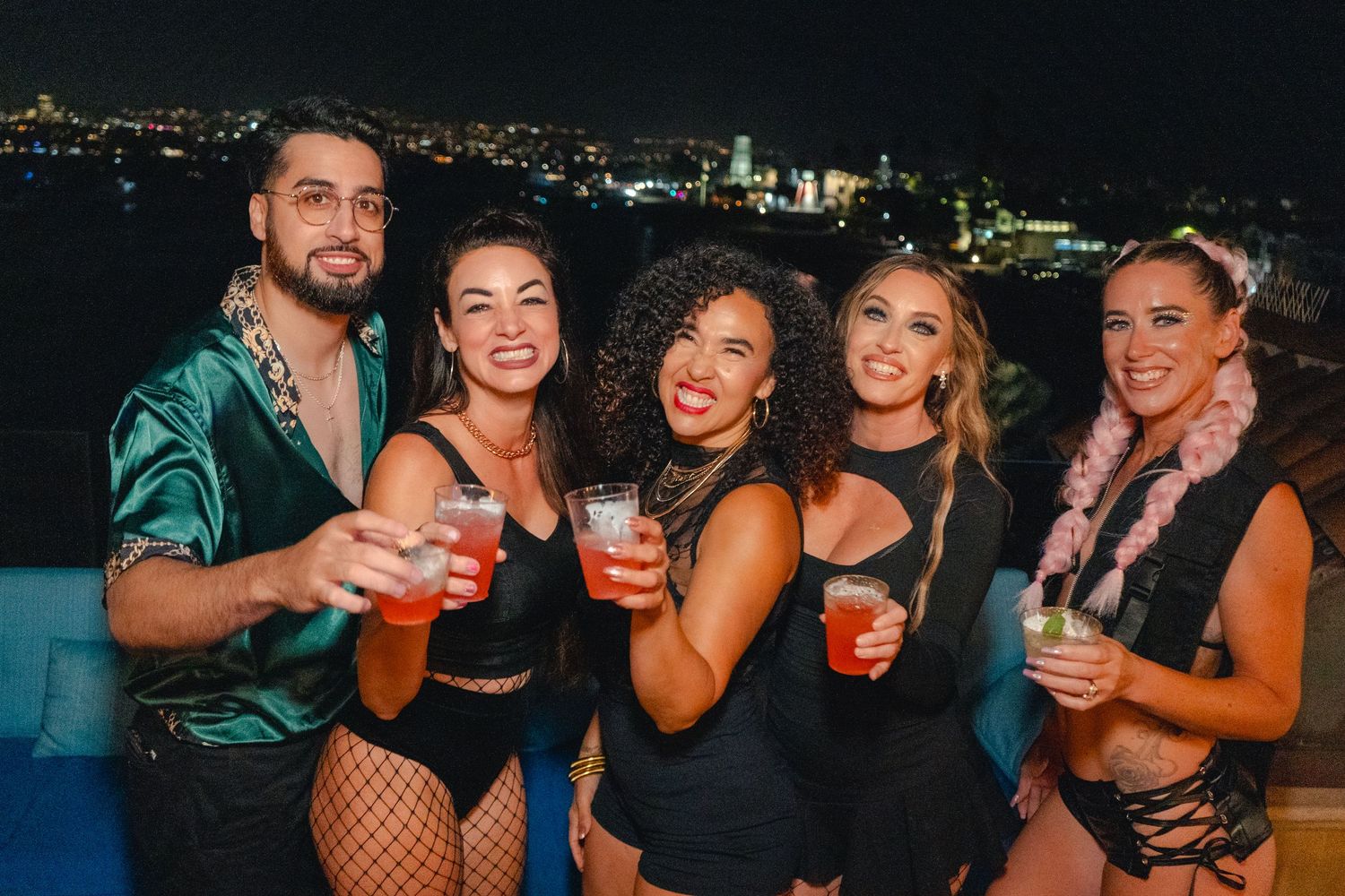 A group of friends enjoying drinks at a rooftop party with a nighttime cityscape visible in the background.