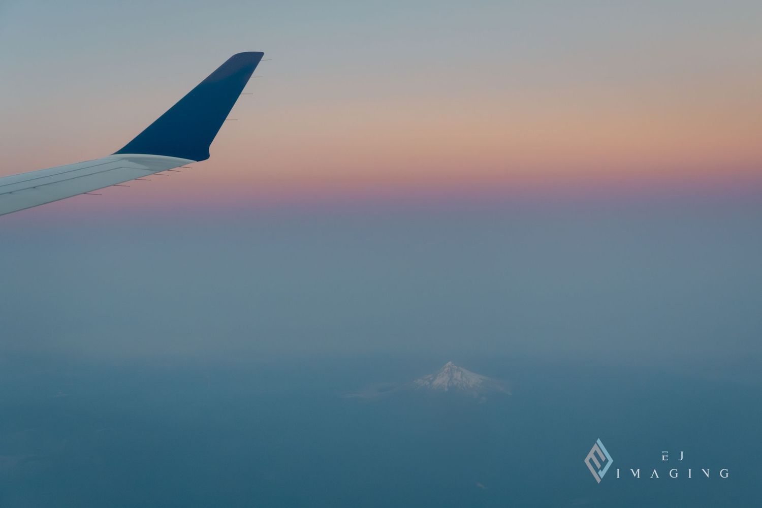 Airplane wing silhouetted against pastel sunset sky with mountain peak visible through clouds below.