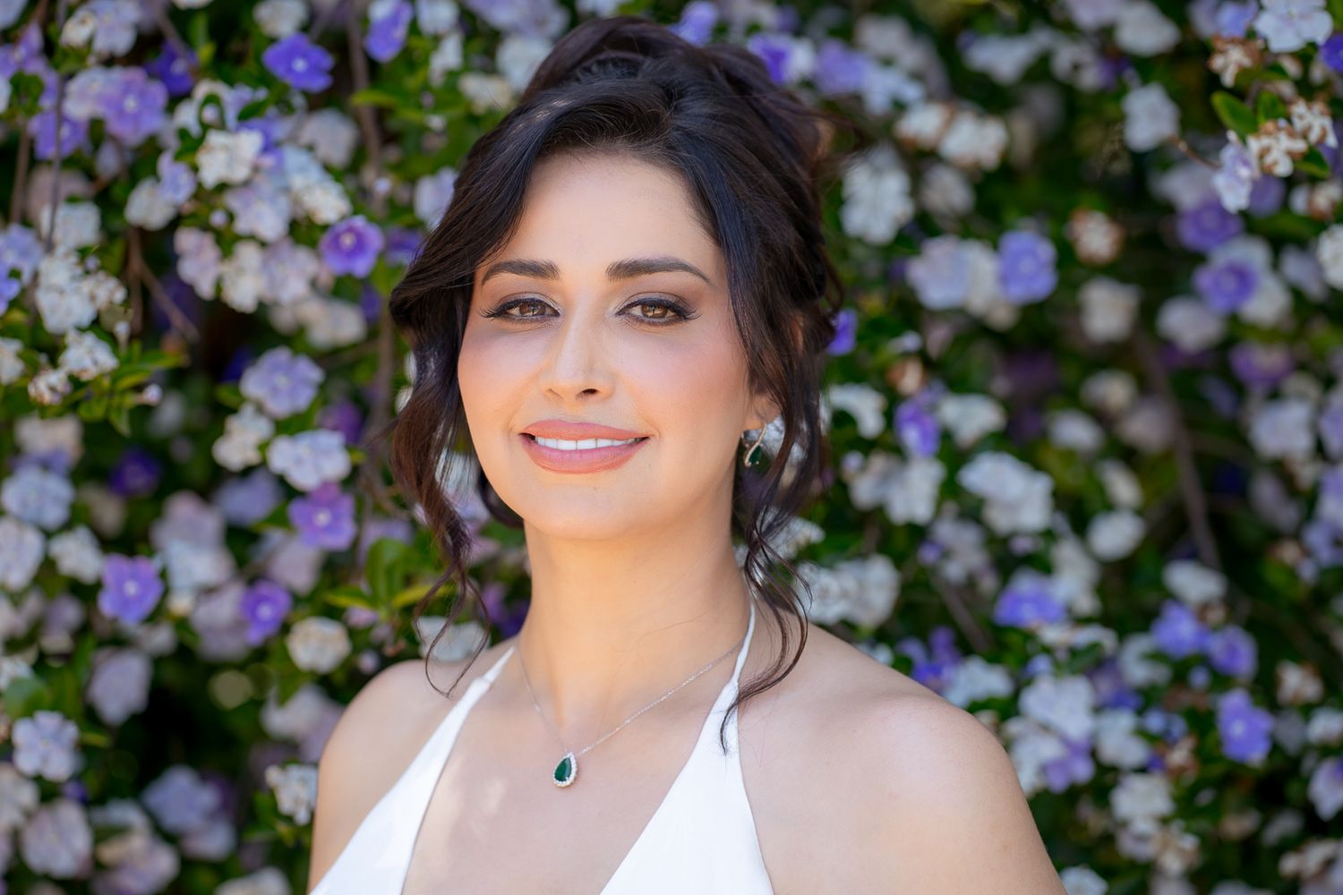 Someone poses in a white dress against a wall of purple and white flowers on a bright sunny day.