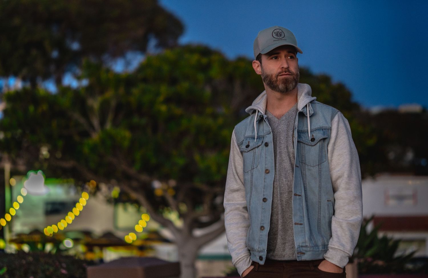 A photographer wearing a denim jacket and cap stands against a blurred outdoor background in evening light.