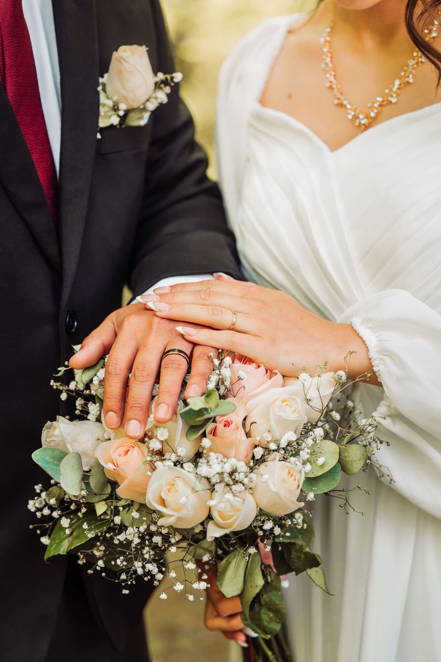 A close-up of newlyweds' hands with wedding rings resting on a peach and white rose bridal bouquet with baby's breath.