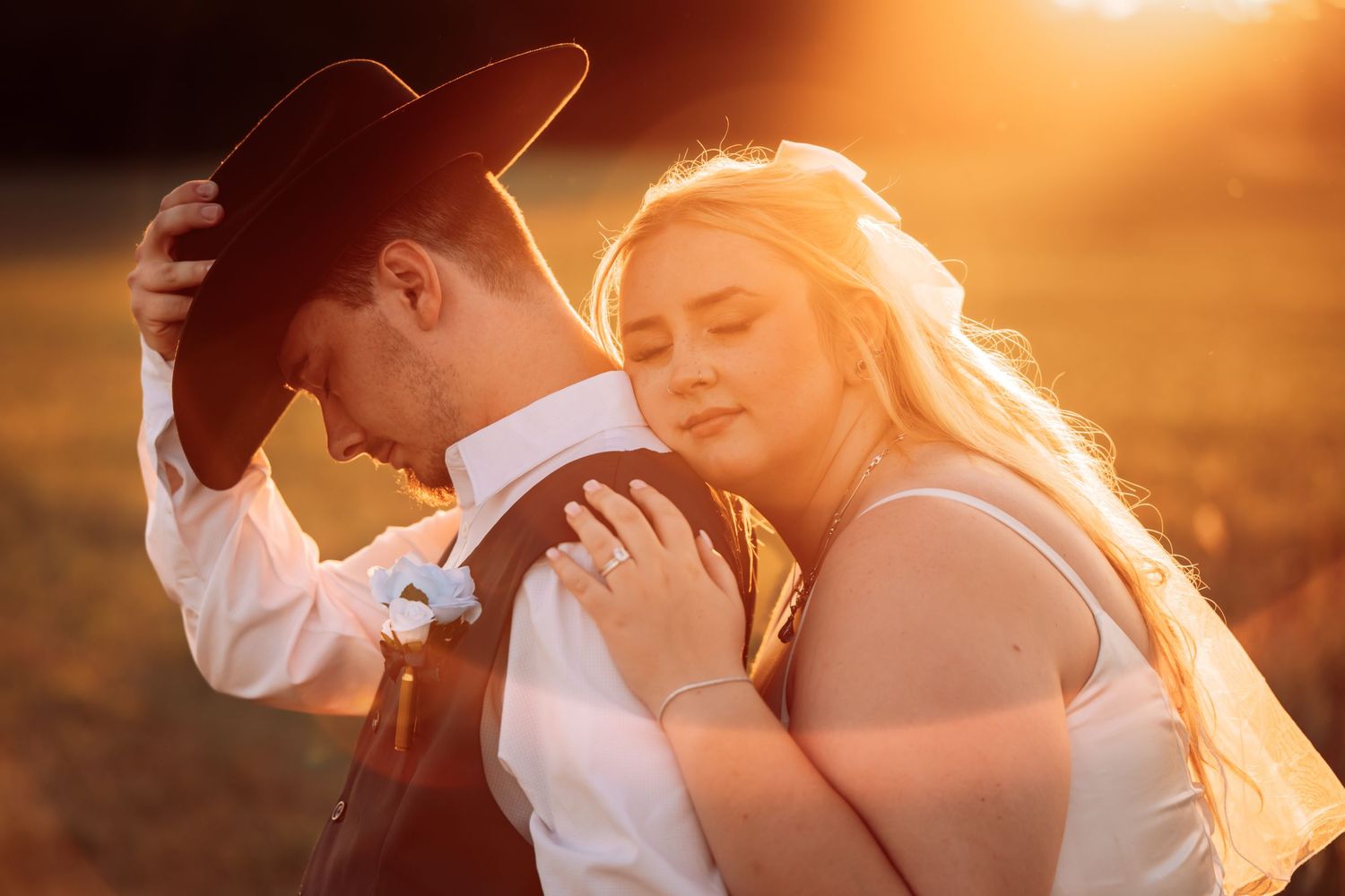 Romantic couple embraces in a field during golden hour sunset as sun creates warm backlit glow around them.