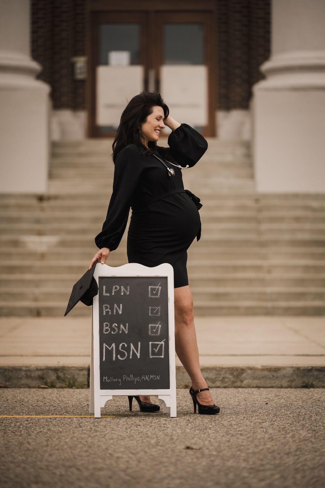 Pregnant woman in black dress stands beside chalkboard sign showing nursing degrees LPN,RN,BSN,MSN all checked off.