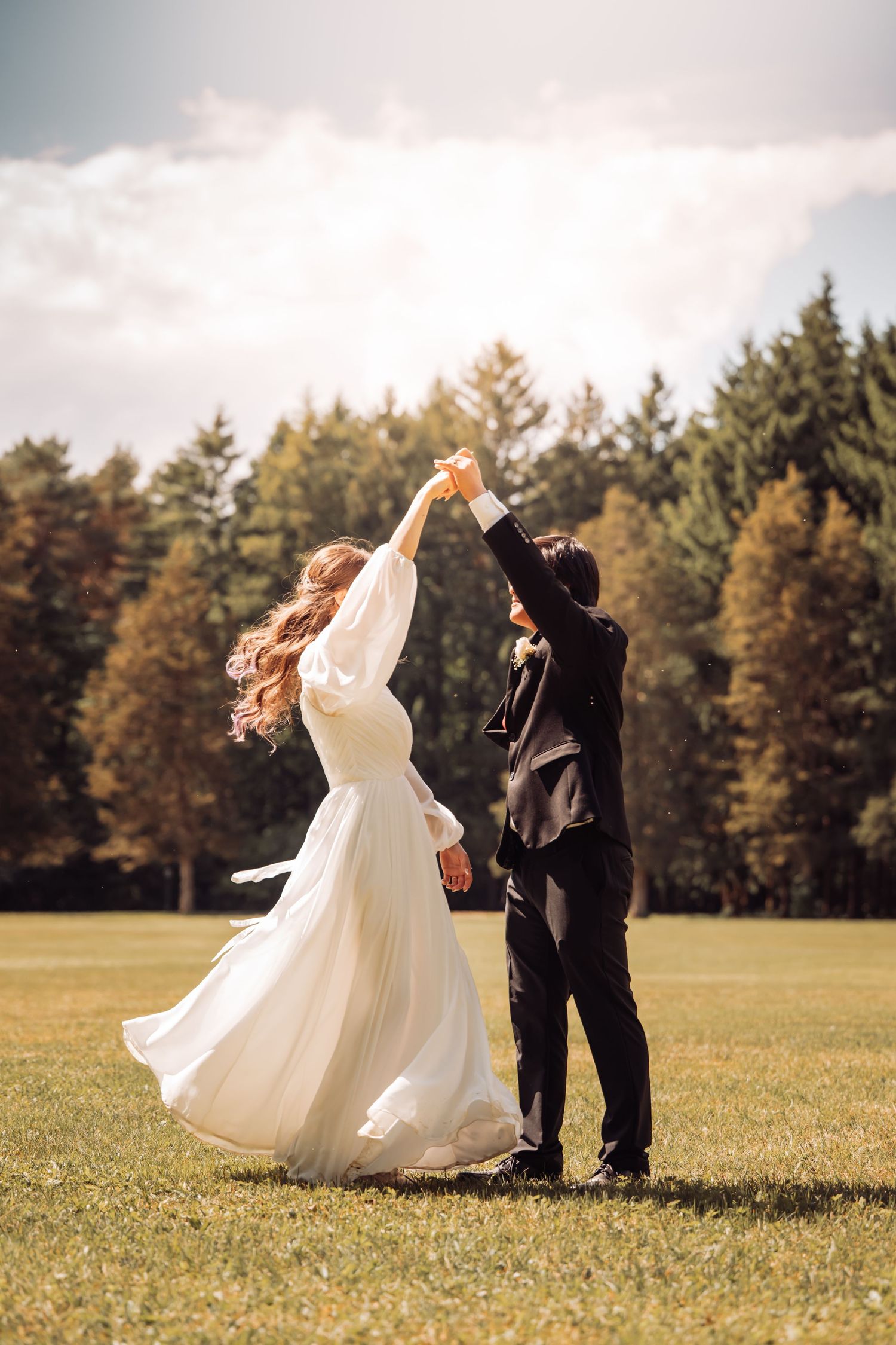 A couple dances in a grassy field with evergreen trees in the background as the bride's white dress flows in the breeze.