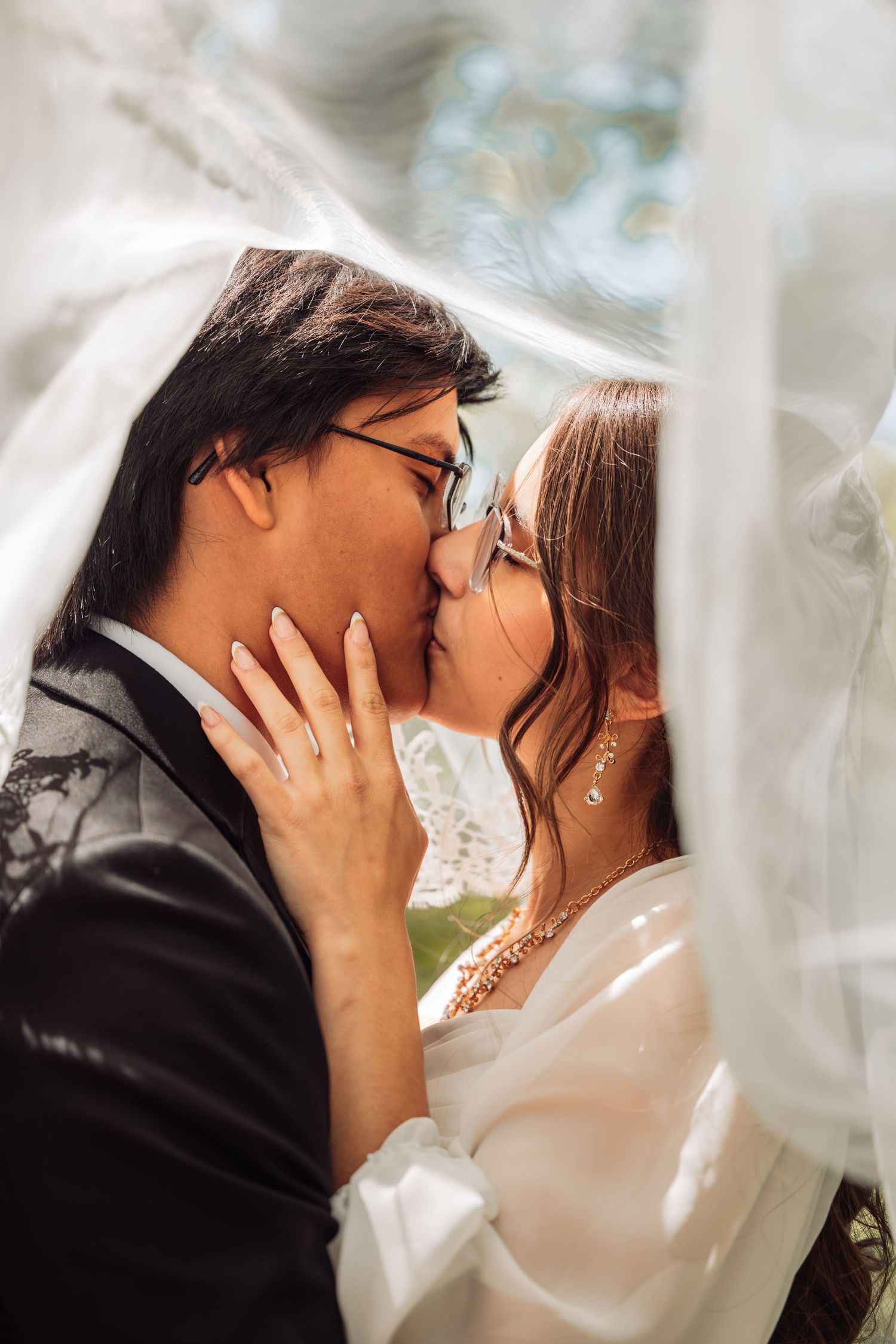 A romantic wedding portrait showing a tender kiss between newlyweds through flowing white veil in soft natural lighting.