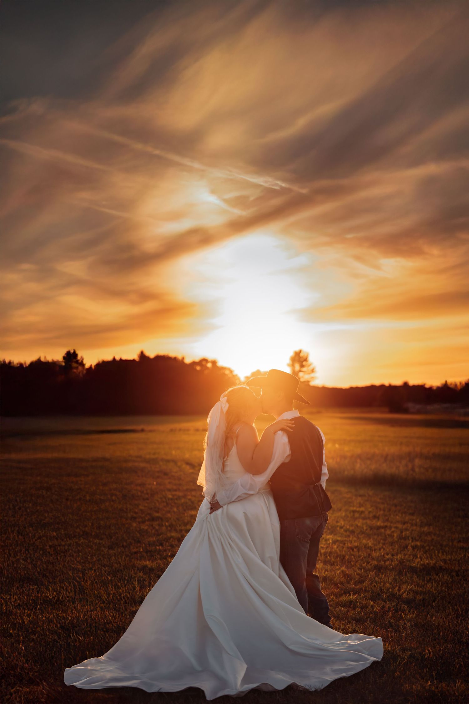 A couple embraces during golden hour sunset in an open field with dramatic orange clouds in the sky.