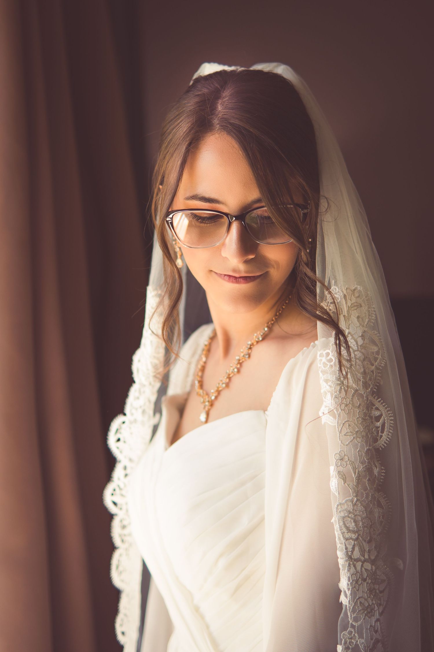 A bride wearing glasses and a lace veil smiles gently while looking down in a soft indoor light.