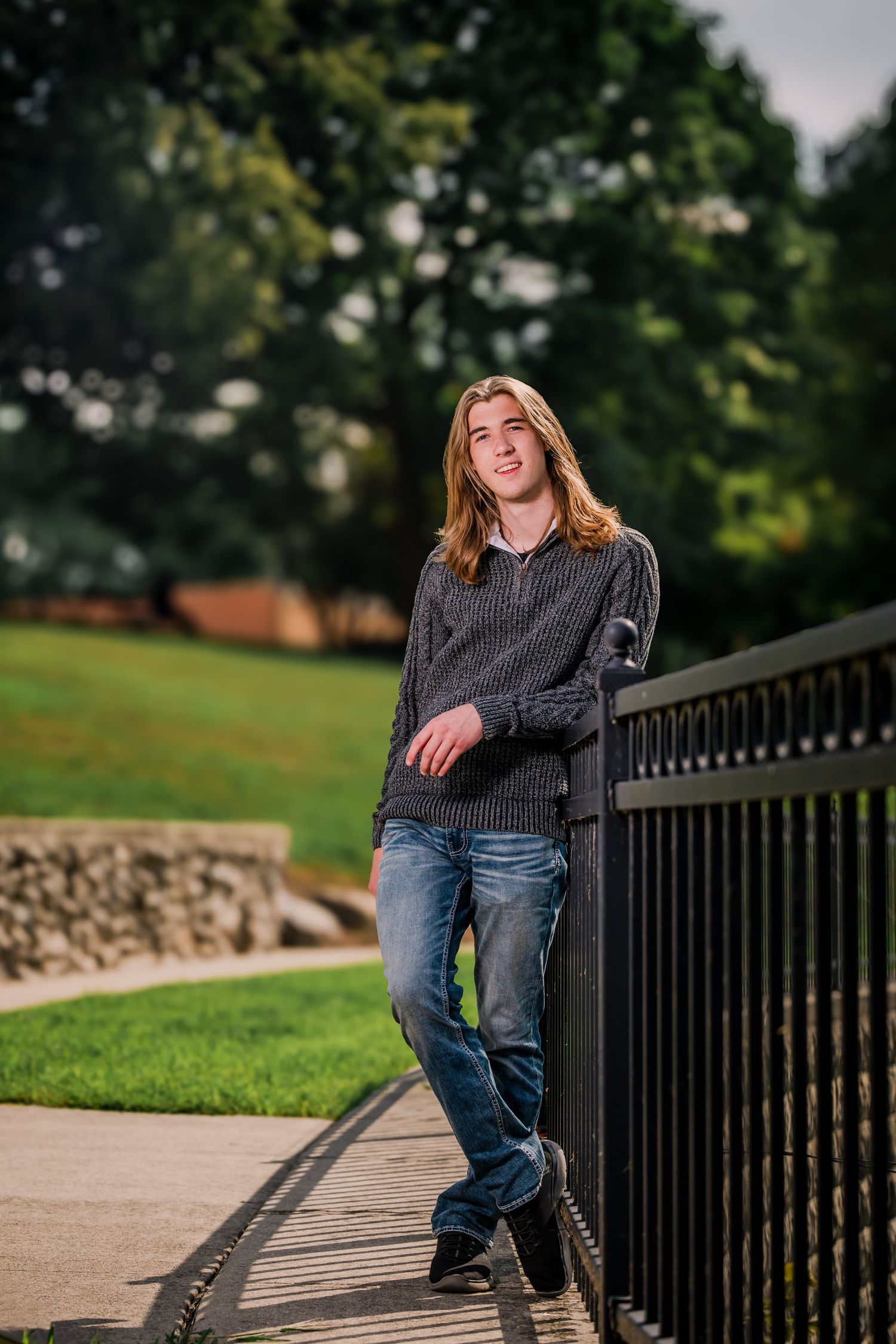 Portrait of someone in a grey sweater and jeans leaning casually against a black iron railing on a path with trees in background.