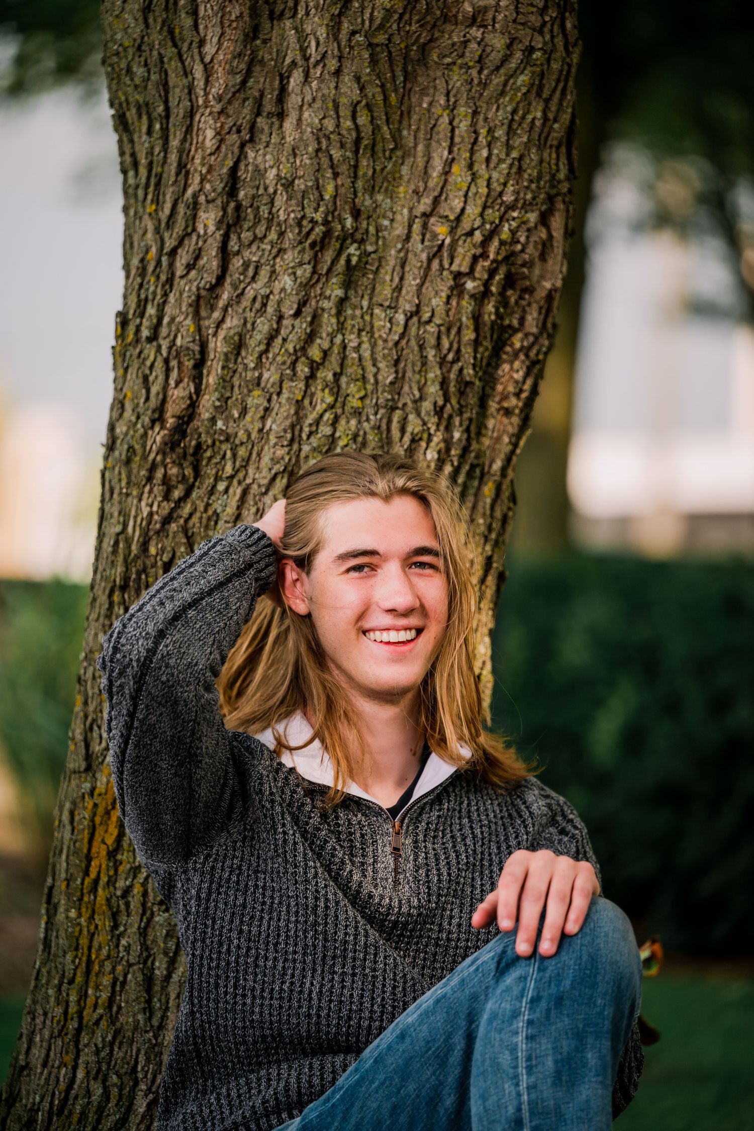 Artistic portrait series showing someone sitting against a tree trunk in a charming outdoor setting wearing a gray sweater.