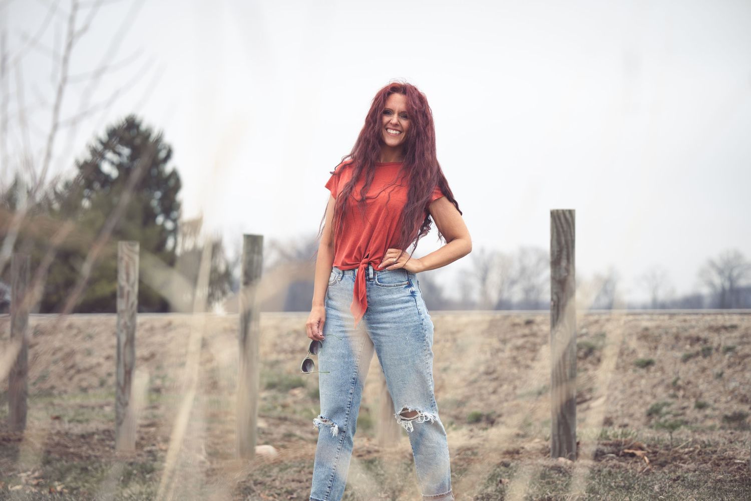 Woman with long red hair in ripped jeans and red top poses confidently outdoors near a rustic wooden fence.