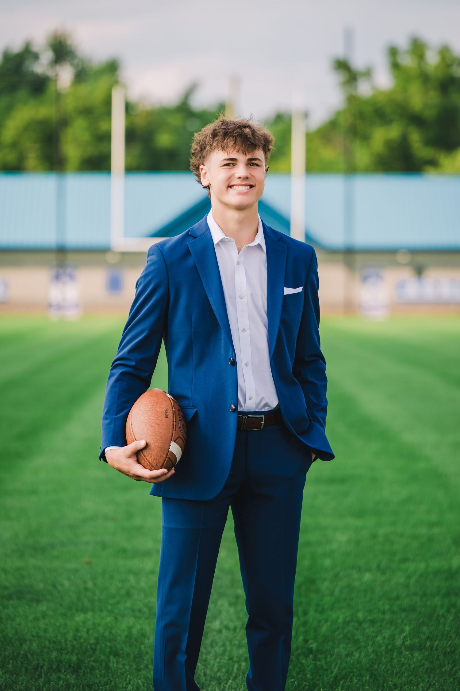 A person in a fitted navy blue suit holds a football while standing on a green sports field.