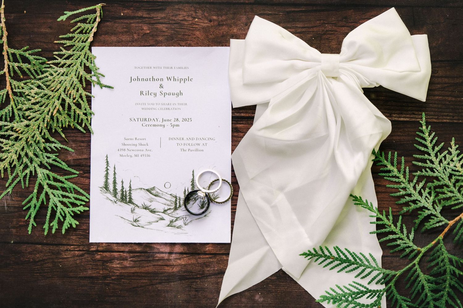 Wedding invitation with pine tree illustration displayed with white bow and evergreen branches on dark wooden surface.