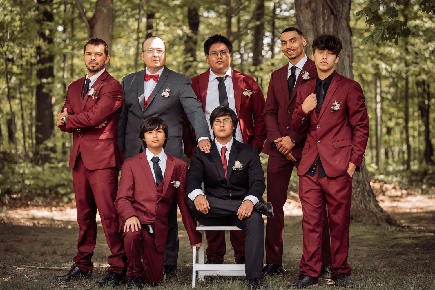 Wedding party wearing matching burgundy suits pose together in a wooded outdoor setting.