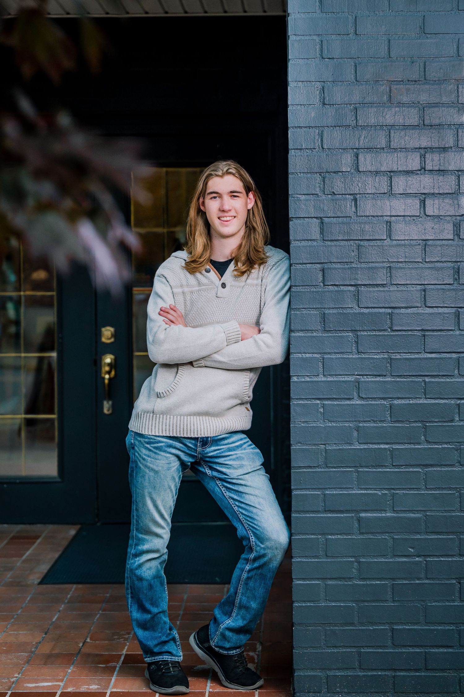 A person in a beige sweater and blue jeans stands confidently against a dark blue brick wall by a doorway.