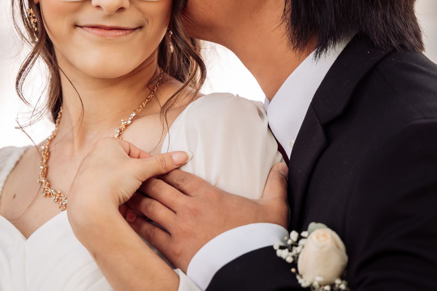 Close-up of newlyweds embracing, with bride in white dress and groom wearing boutonniere and black suit.