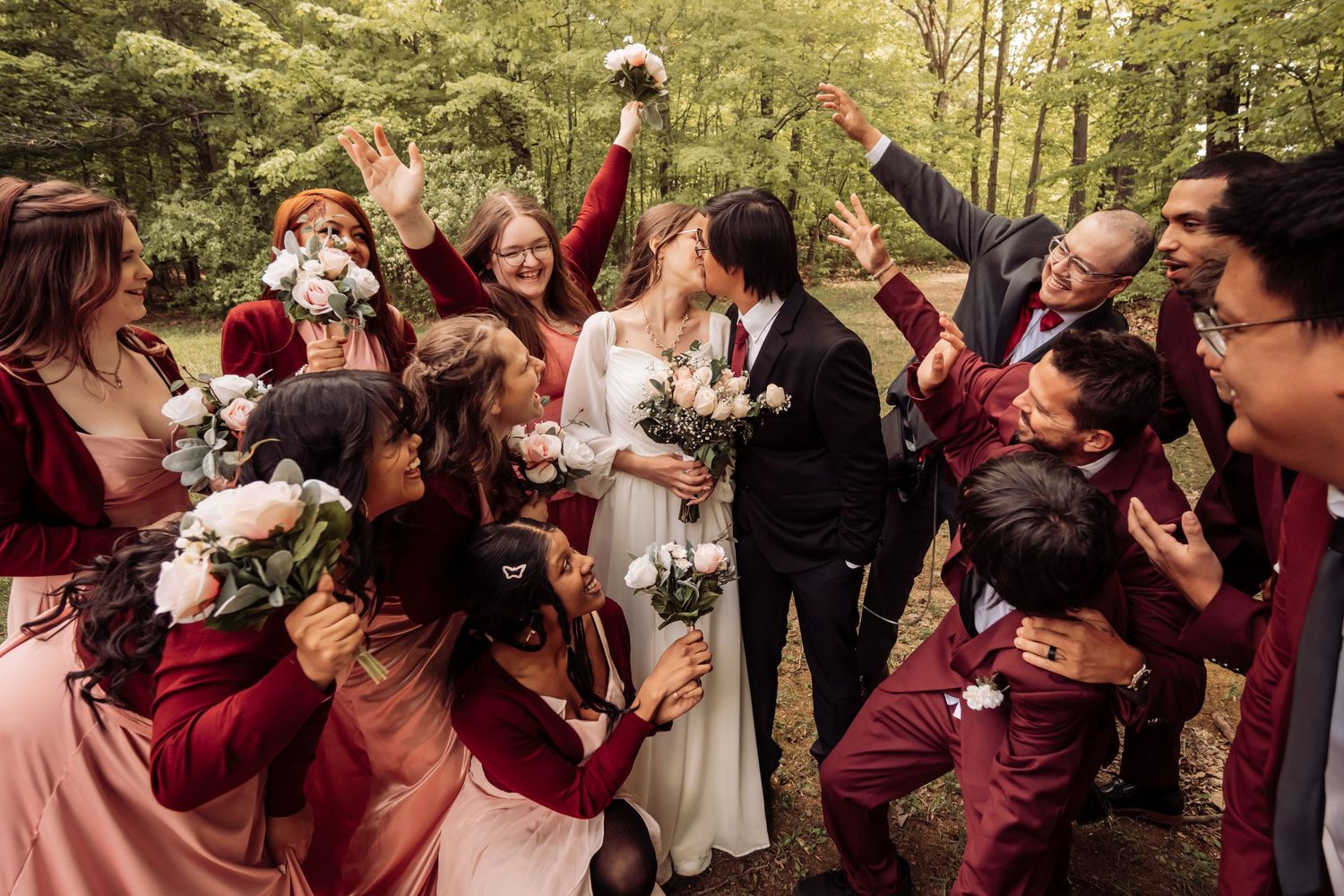 Wedding party celebrates outdoors with joyful cheers and raised arms while wearing burgundy attire.