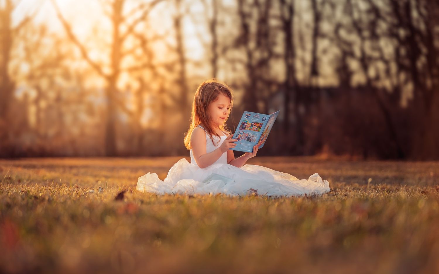 Young girl in white dress sitting on grass reading a book outdoors in golden sunset light.