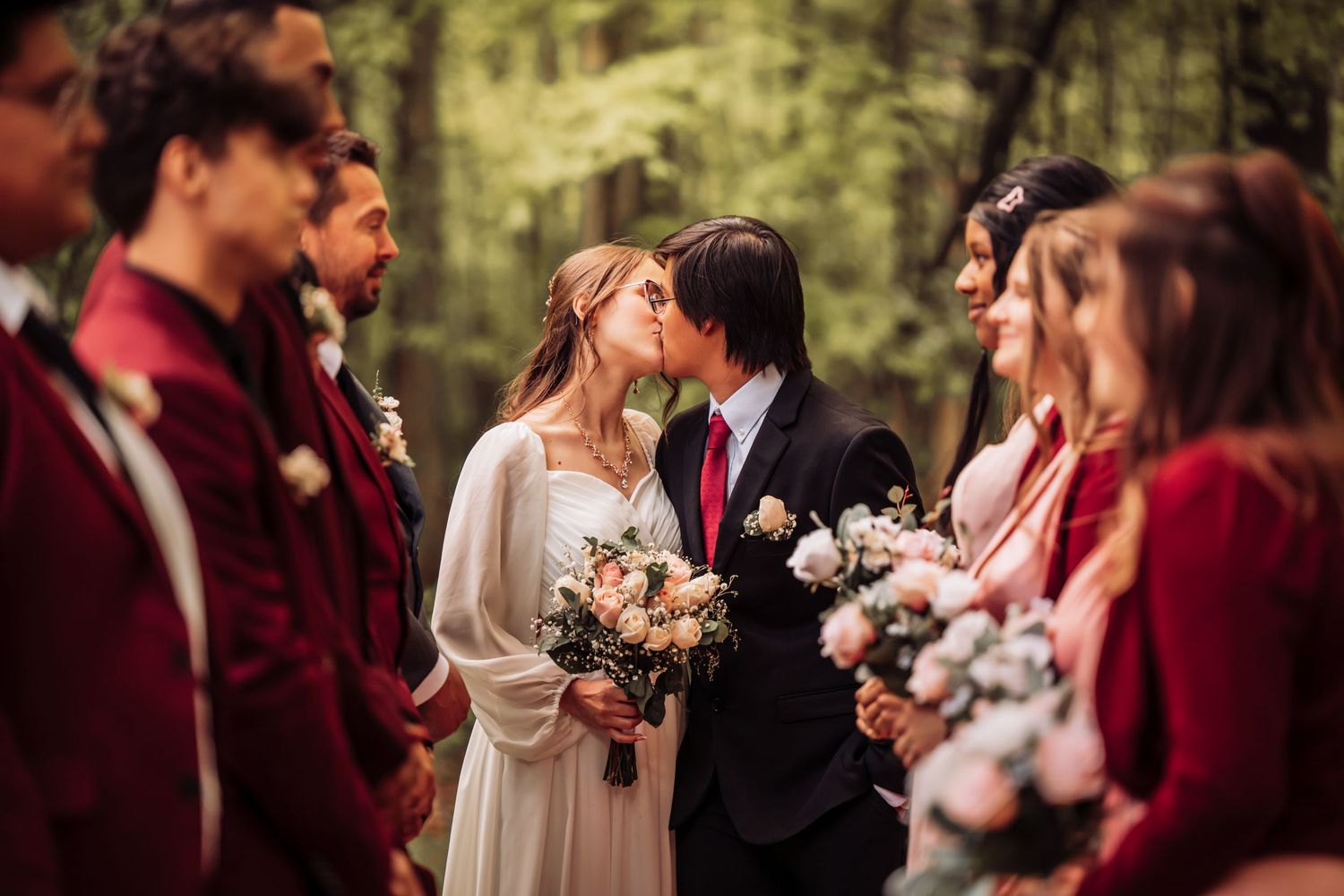 Couple shares a romantic moment surrounded by wedding party members in burgundy attire at outdoor forest ceremony.