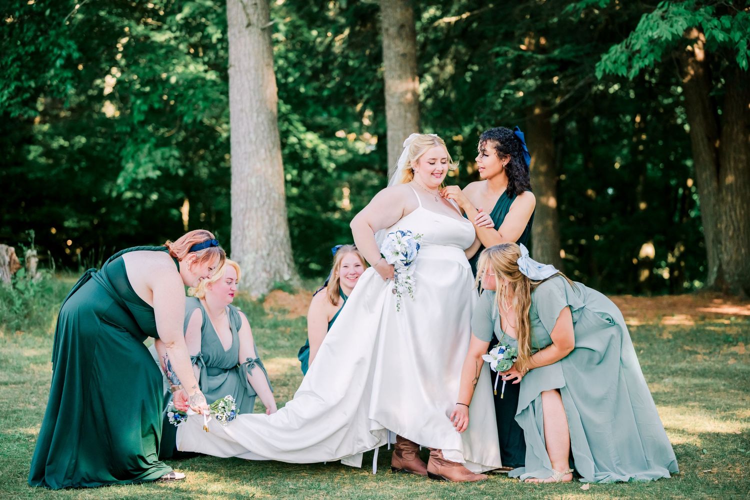 Bridesmaids in teal and blue dresses help adjust a flowing white wedding gown in a sunlit forest setting.