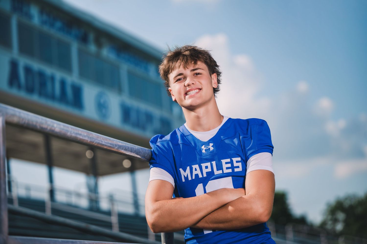 A high school football player in blue and white uniform stands confidently on the field during a sunny day.