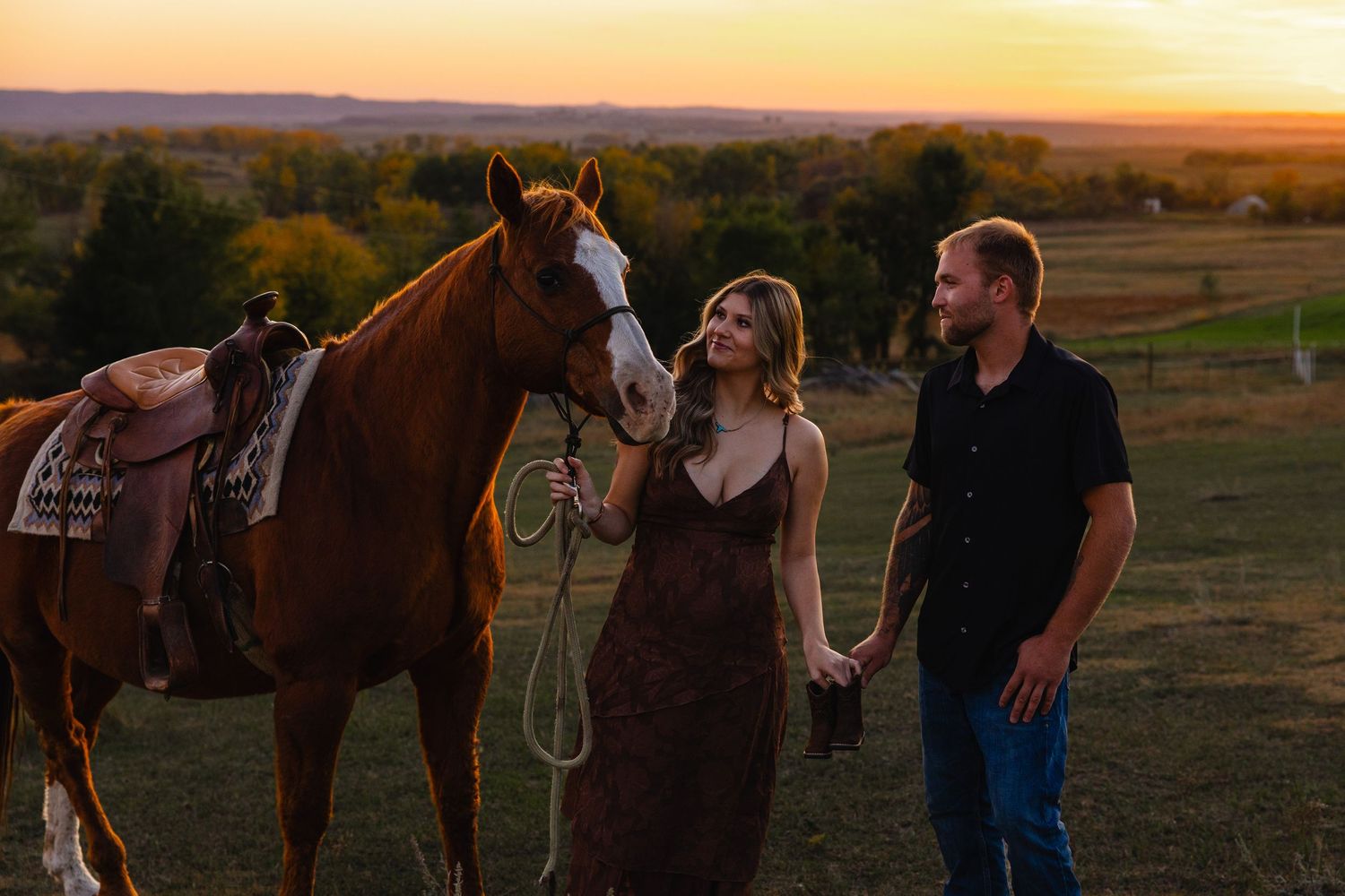 Couple holding hands at sunset with a saddled chestnut horse in a scenic countryside field.