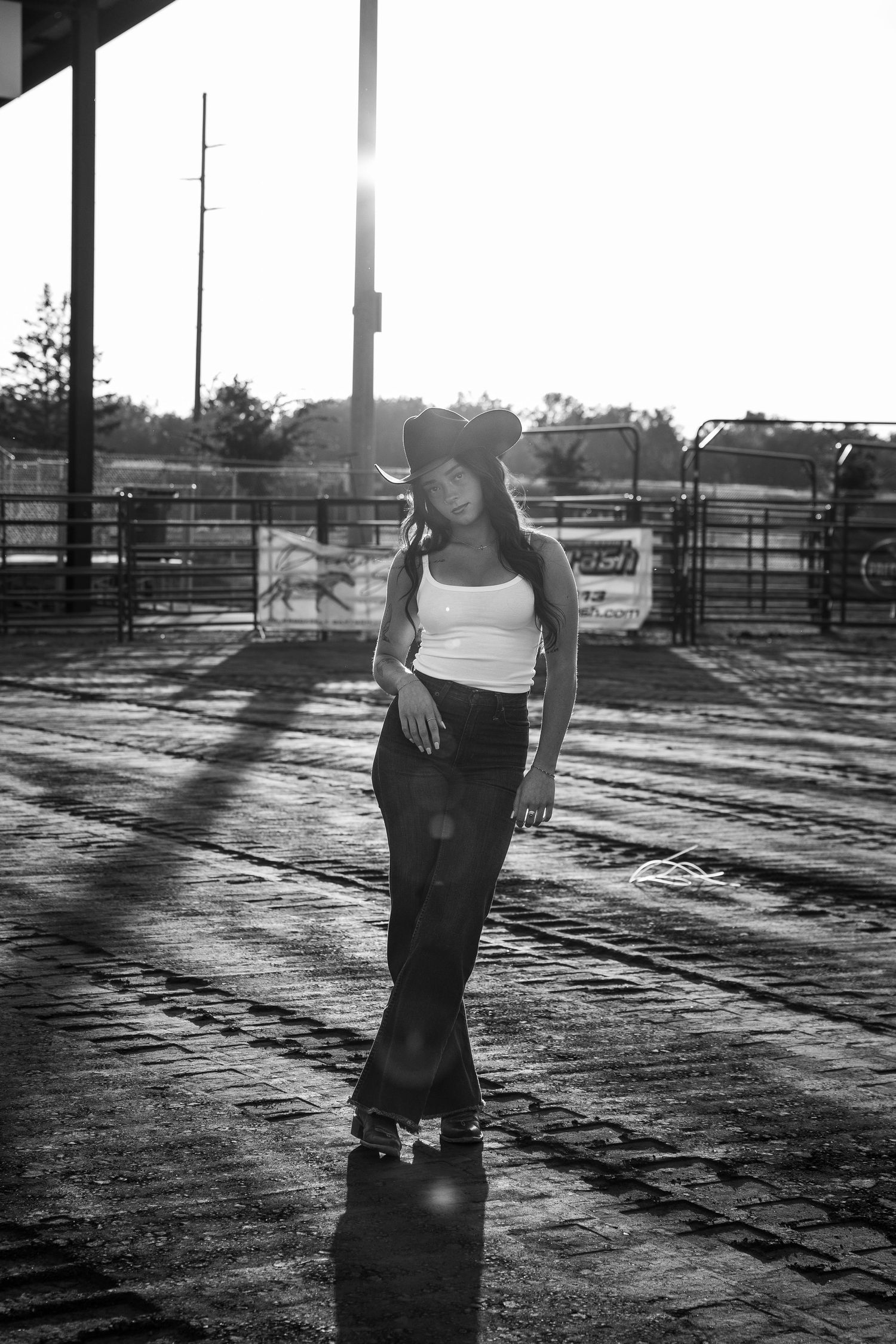 Black and white portrait of a woman in a cowboy hat and tank top standing in a sunlit rodeo arena with fences and pens behind her.