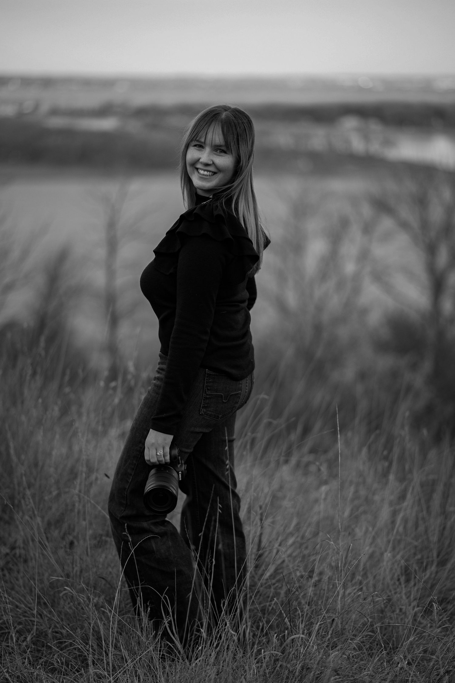 Black and white portrait of smiling woman holding camera in field.