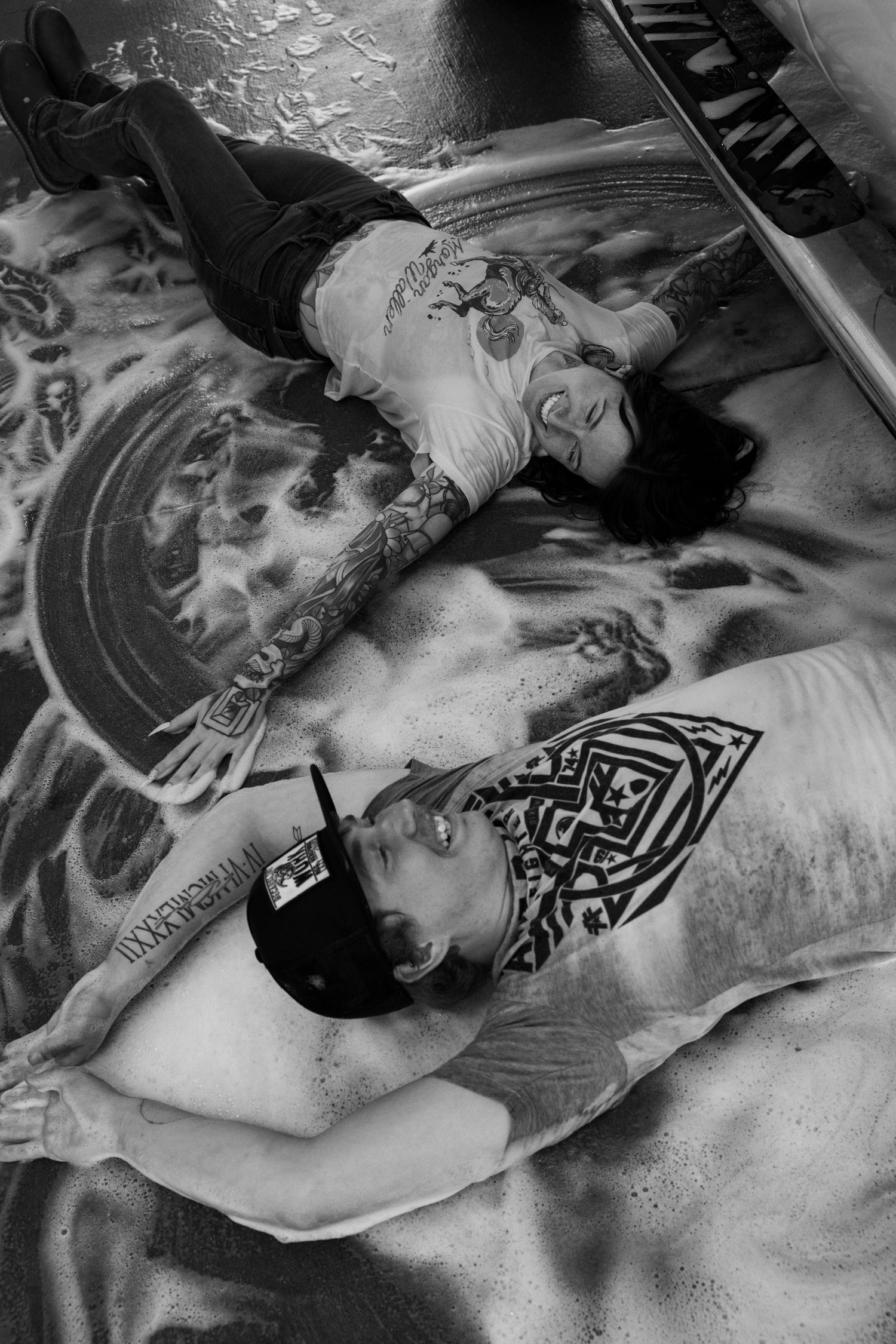 Black and white photo of two tattooed people lying on a reflective wet surface.