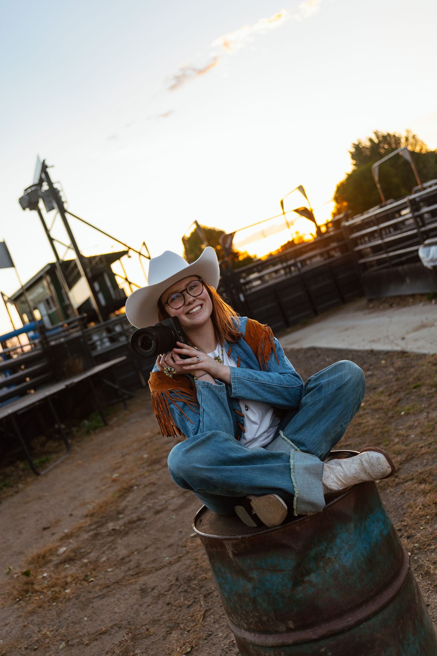 Smiling woman in cowboy hat and denim jacket sitting cross-legged on barrel at ranch during golden hour sunset.