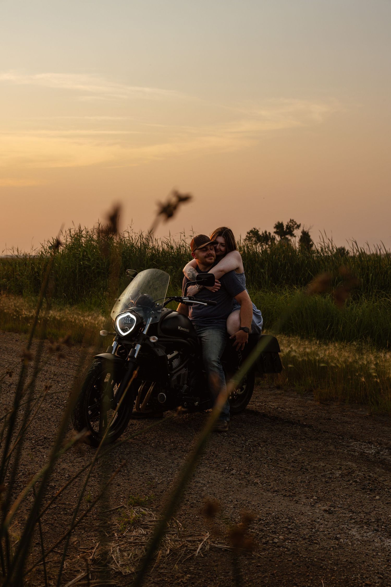 Couple embracing on a motorcycle on a rural dirt road at sunset.