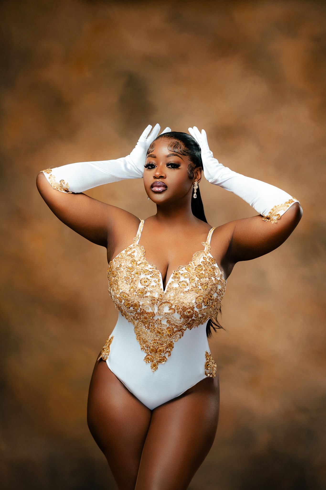 Glamour portrait in white and gold bodysuit with matching gloves against brown backdrop.
