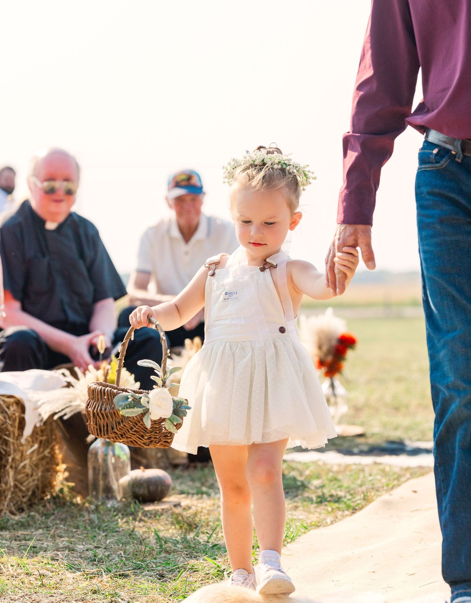 A small child in a white dress walks along a path holding flowers at an outdoor event.