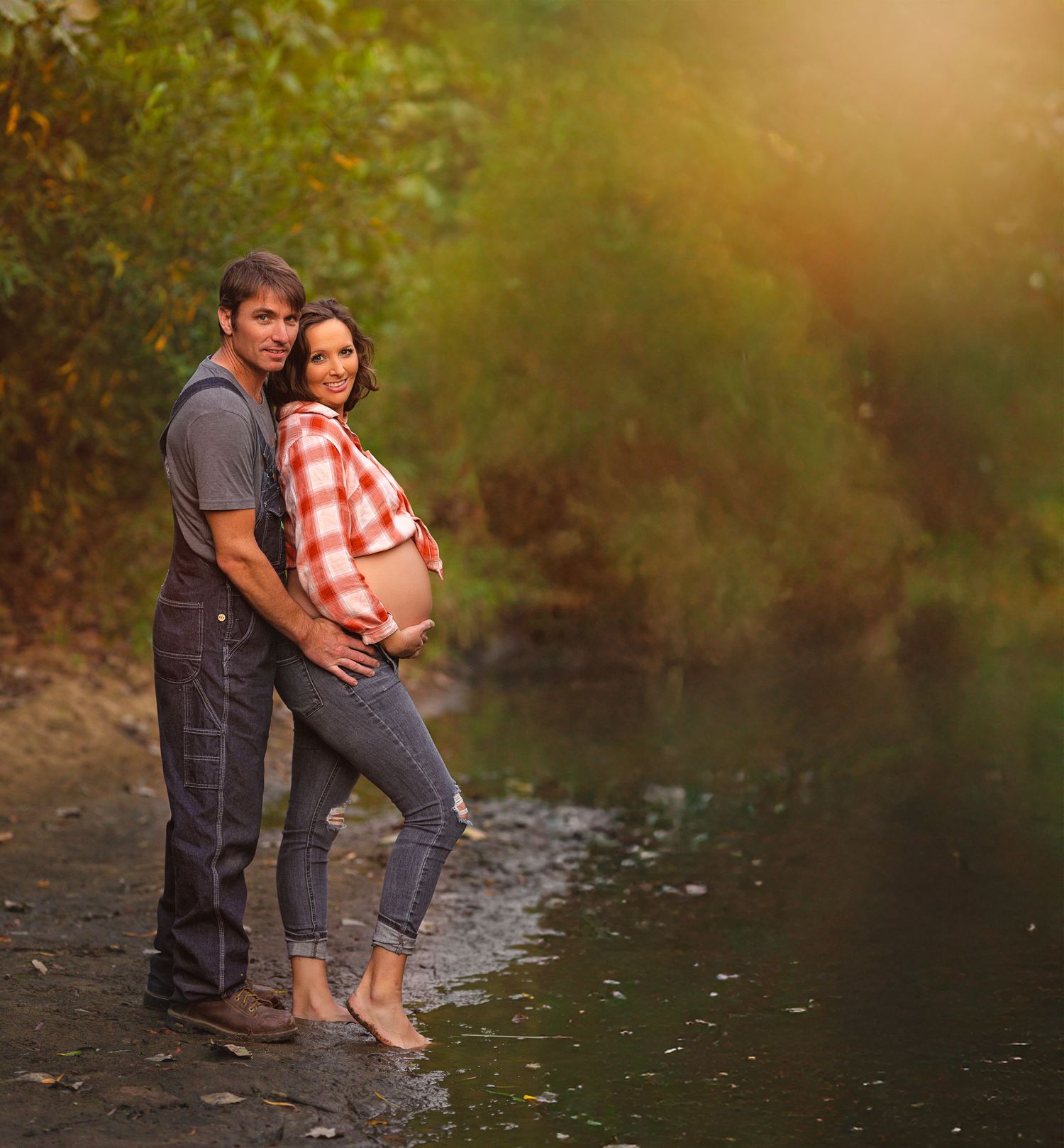 Couple poses for maternity photo by a creek at sunset in casual attire.