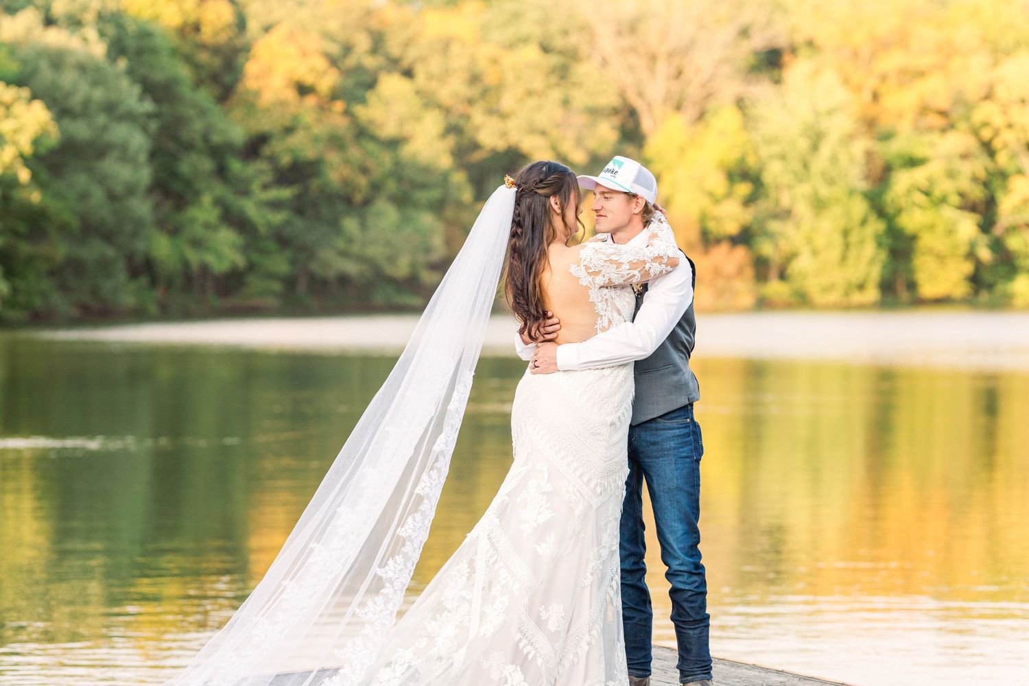 Couple embracing by a lake with flowing wedding veil in golden sunset light.
