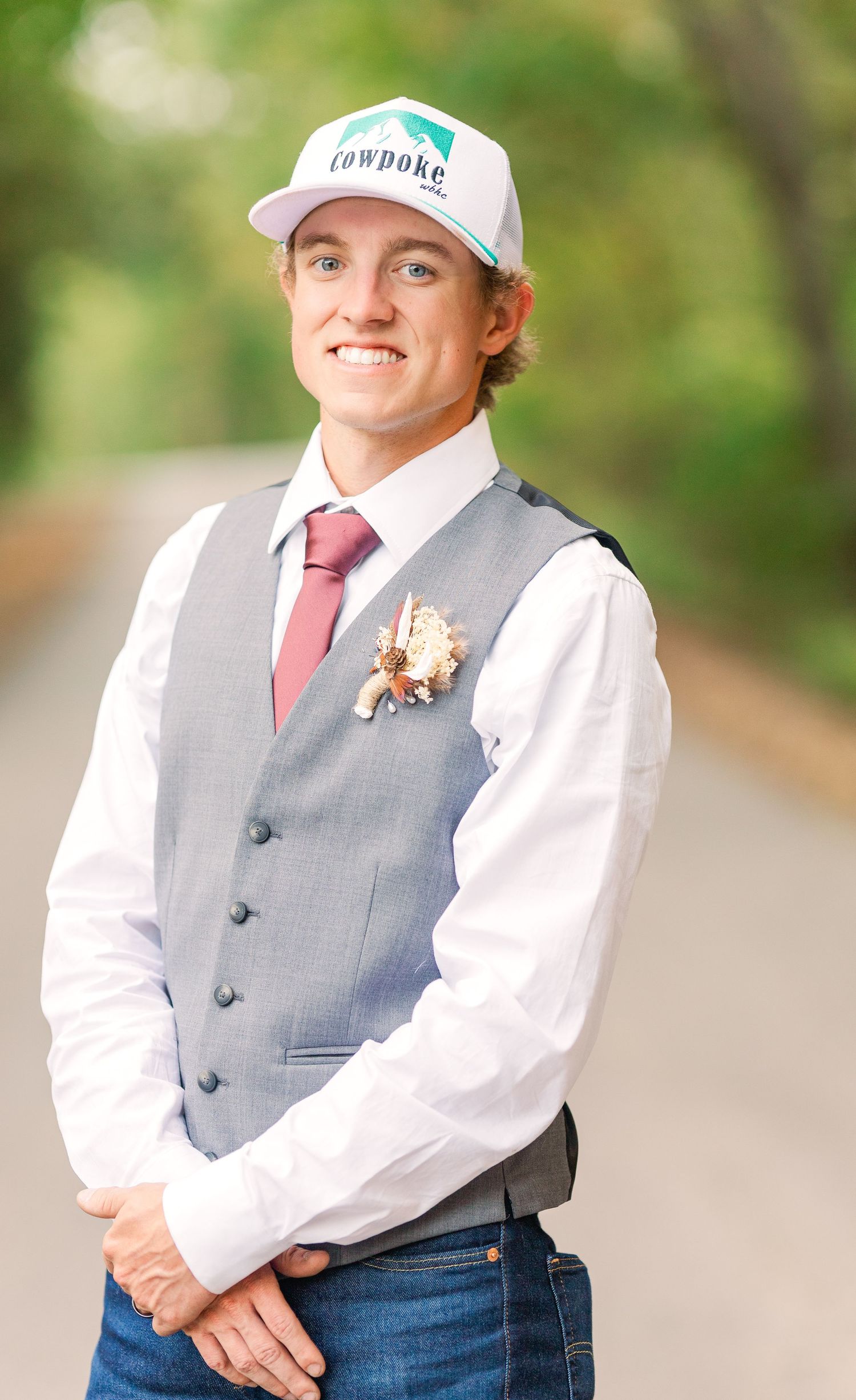 Groom in casual attire with grey vest and baseball cap against outdoor backdrop.