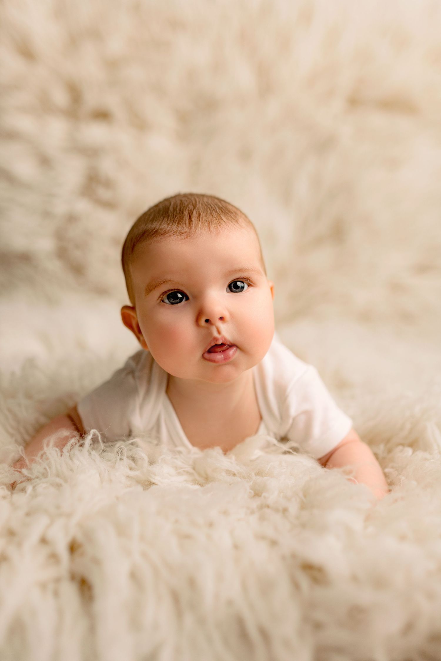Baby in white outfit lying on soft furry blanket during studio photoshoot.