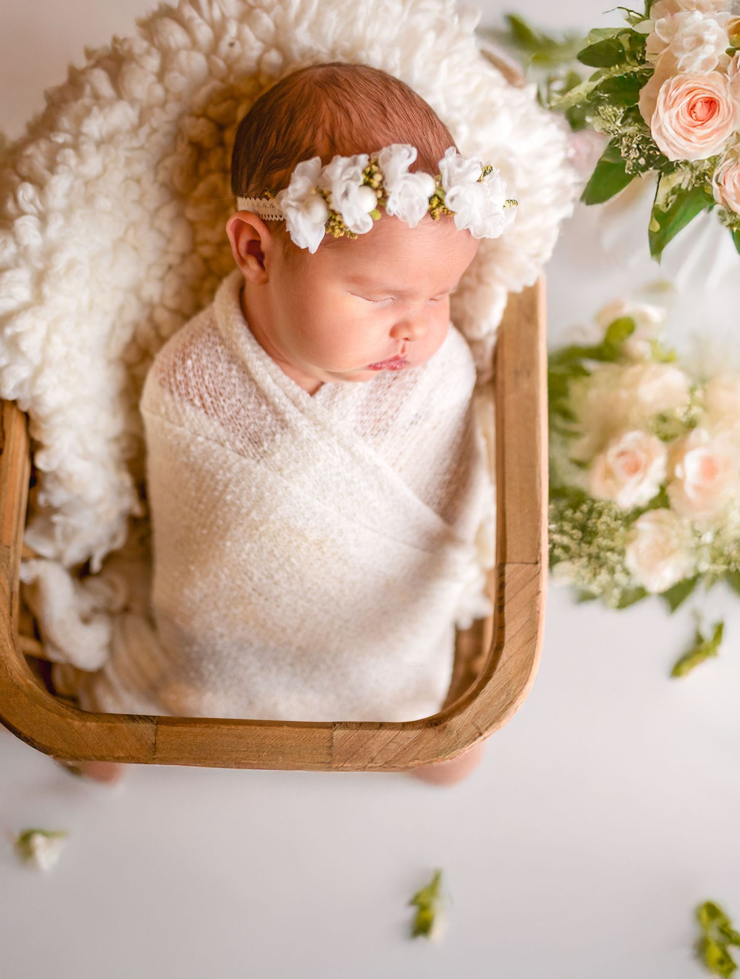 Newborn sleeping in wooden cradle surrounded by roses and wearing floral crown.